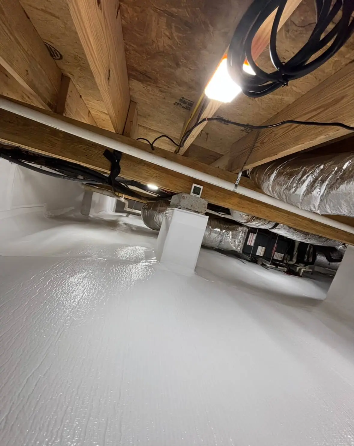 A bright, wide view of a fully encapsulated crawl space showing the white vapor barrier on the floor and walls, a small dehumidifier unit and drain pipe in the back, and a window opening on the right wall.
