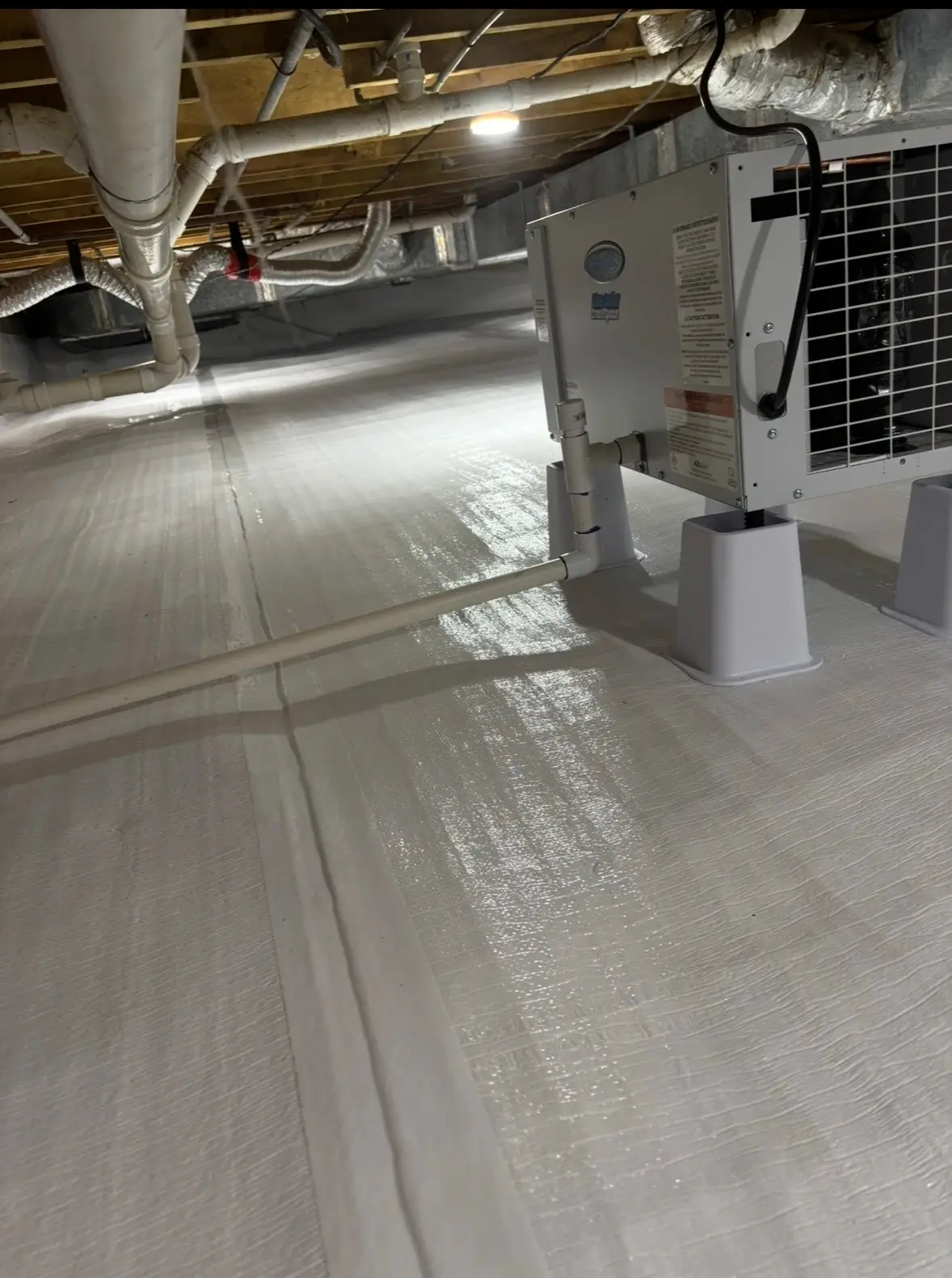 Crawl space dehumidifier with condensate line on a white vapor barrier beside insulated ductwork